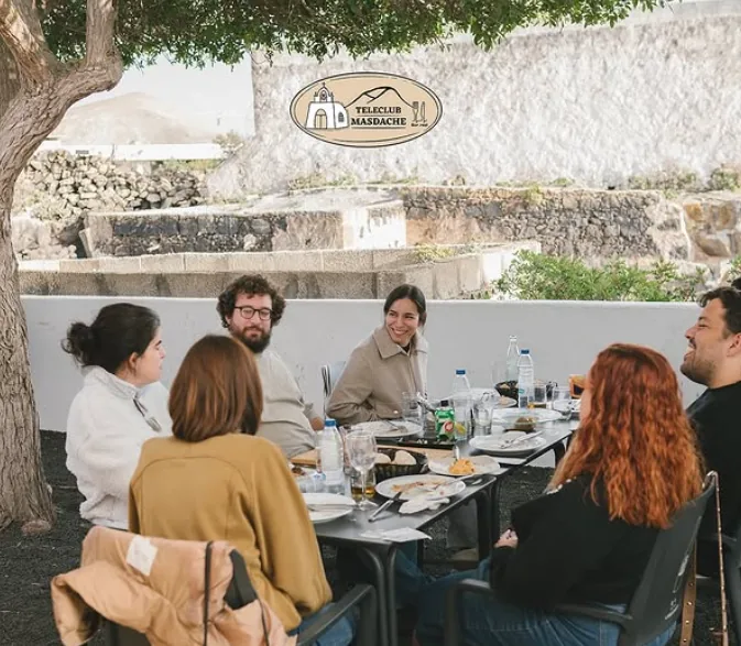 Una mesa larga con amigos comiendo en la terraza del Teleclub Masdache, junto al muro de piedra volcánica
