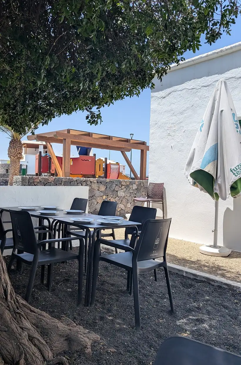 Terraza del Teleclub Masdache con mesas puestas bajo el árbol, muro de piedra volcánica y el parque infantil detrás